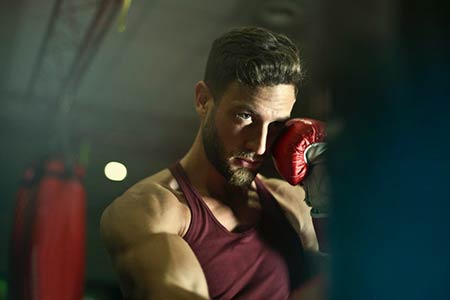A boxer with a beard wearing a red top punches a heavy bag.