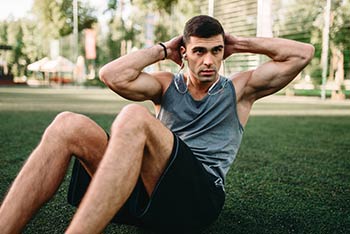 A male athlete doing some sit-ups trains outdoors on grass.