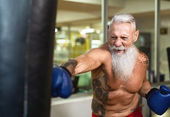 A strong elderly man hits the punch bag in the gym.